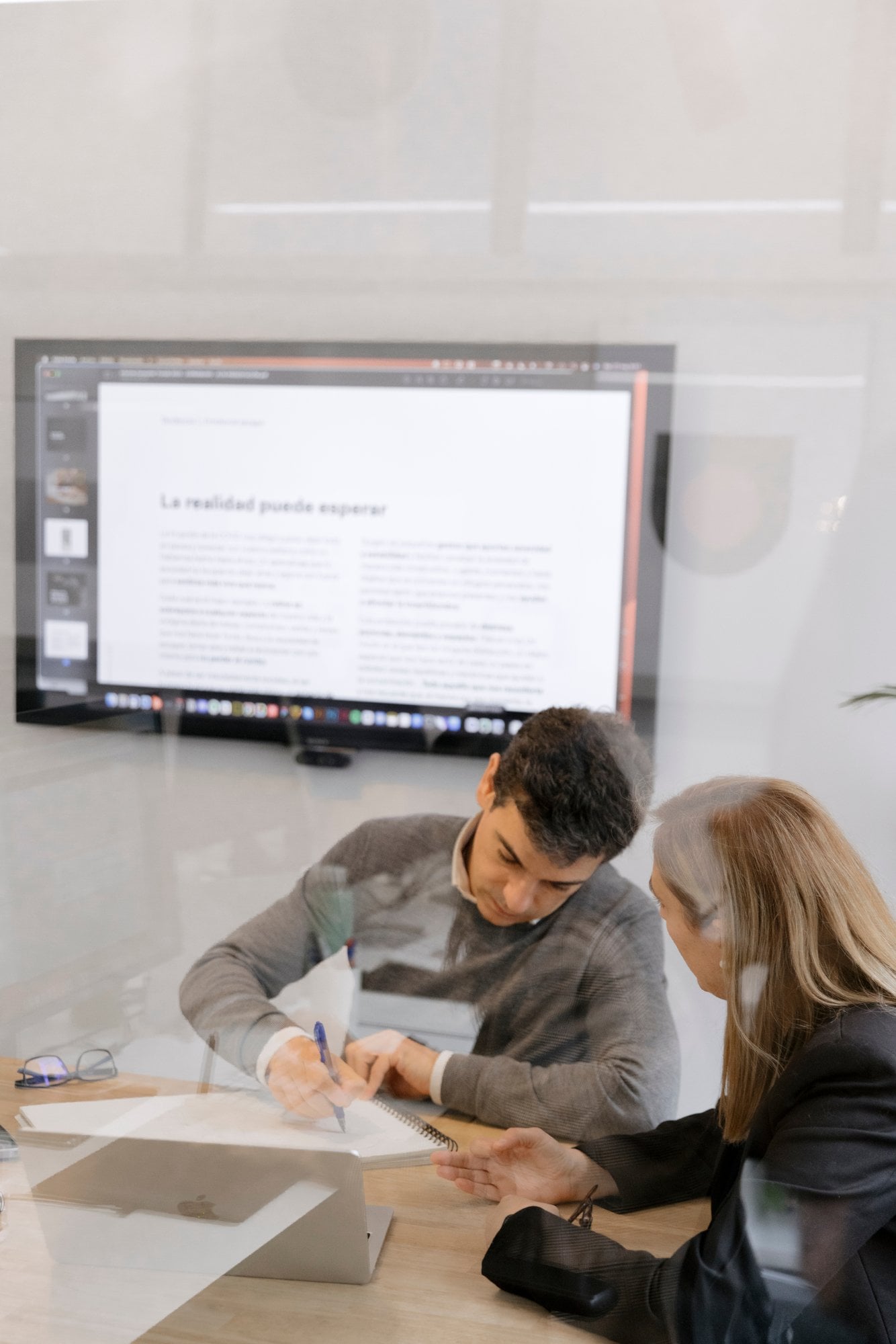 Dos compañeros revisando documentos frente a una pantalla, tras un cristal